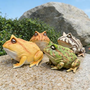 Może przedstawiać: Cztery ceramiczne figurki żab w różnych kolorach: żółtym, pomarańczowym, zielonym i brązowym z białymi i brązowymi paskami. Żaby są ustawione na kamiennej powierzchni, z dużą skałą i zielenią w tle.