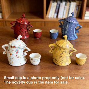 以下が含まれることがあります： 赤、青、白、黄色の蓋付きセラミックノベルティカップ4個。各カップはユニークなデザインで、小さなカップと組み合わされています。画像には「小さなカップは写真撮影用のみ（販売していません）。ノベルティカップが販売商品です」と書かれています。