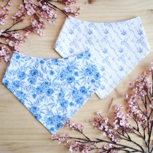 May include: Two white bibs with blue floral and ribbon patterns. One bib features a dense pattern of blue roses, while the other has a delicate ribbon and floral design. The bibs are displayed on a wooden surface with pink floral accents.
