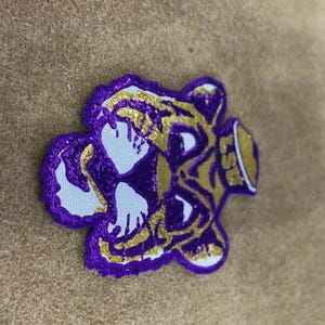 May include: Embroidered patch with a purple and gold tiger head design. The patch has a white background and the letters "LSU" are visible. The patch is likely for apparel or accessories.