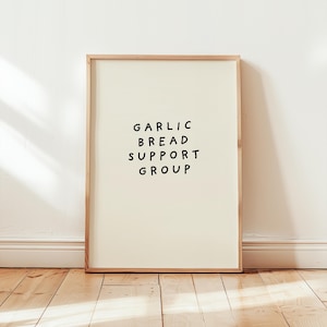 May include: A rectangular wall art print in a light wood frame featuring a cream-colored background with the black hand-lettered text GARLIC BREAD SUPPORT GROUP centered in the middle of the print in a simple sans-serif font style.