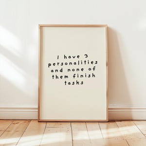 May include: A rectangular wall art print in a light wood frame featuring a cream-colored background with black handwritten-style text that reads: I have 3 personalities and none of them finish tasks. The print is centered within the frame.