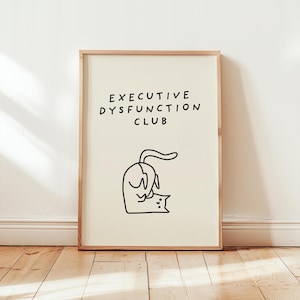 May include: Framed art print with the text "EXECUTIVE DYSFUNCTION CLUB" above a whimsical black and white illustration of a cat and a dog. The artwork is displayed on a cream-colored background.