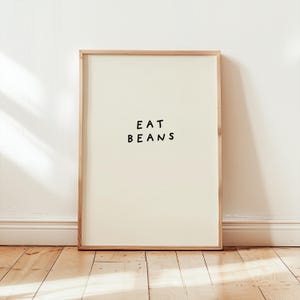 May include: A framed art print with the words "EAT BEANS" in black, centered on a cream-colored background. The frame is a light wood color, and the print is leaning against a white wall and wooden floor.