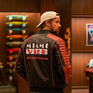 Puede incluir: Chaqueta de cuero negra y roja con el texto "MIAMI VICE STUNTTEAM" en la espalda. La chaqueta tiene paneles acolchados rojos en los hombros y un cuerpo negro con ribetes rojos. La persona lleva una gorra de béisbol blanca y vaqueros azules.
