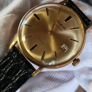 Könnte beinhalten: Eine vintage goldfarbene Glashütte Pezimatic Armbanduhr mit schwarzem Lederarmband. Das Zifferblatt der Uhr ist goldfarben mit schwarzen Markierungen und Zeigern. Sie hat eine Datumsanzeige bei 3 Uhr. Eine klassische Uhr.