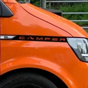 May include: A vibrant orange camper van with black detailing. The word "CAMPER" is displayed in black lettering on the side. The front headlight is visible.