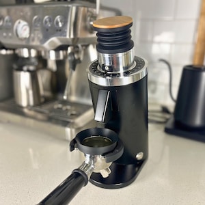 May include: A white ceramic mug with a black handle and the text "But First, Coffee" in a bold, black font. The mug is set against a blurred background, suggesting a cosy setting.