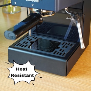 May include: A black espresso machine featuring a portafilter, a drip tray, and a steam wand. A small digital scale is positioned on the drip tray. A white speech bubble graphic with black text reads Heat Resistant, pointing toward the machine base.