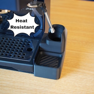 Könnte beinhalten: Ein schwarzes, hitzebeständiges Silikon-Zubehör für eine Espressomaschine mit einer runden Halterung für eine Dampfdüse und einer rechteckigen Abtropfschale mit diagonalen Rillen. Eine Sprechblase mit dem Text Heat Resistant ist zu sehen.