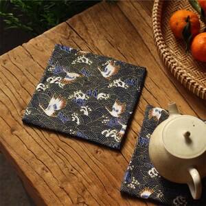 May include: Square fabric coasters with a Japanese wave and crane design. The coasters feature a navy blue background with gold, white, blue, and orange accents. A teapot and oranges in a basket are also visible.