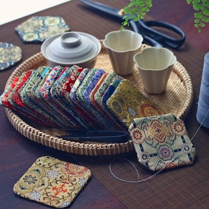 May include: A collection of colorful, patterned fabric coasters arranged on a woven tray. The coasters feature various floral and geometric designs in shades of red, green, blue, and gold. Other items include a ceramic tea set, scissors, and small cups.