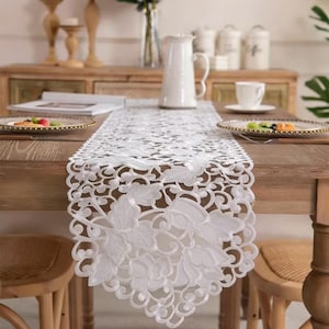 May include: A white lace table runner with a floral design is laid on a wooden table. The table is set with plates, a pitcher, and a vase of flowers. Two wooden chairs are positioned on either side of the table.