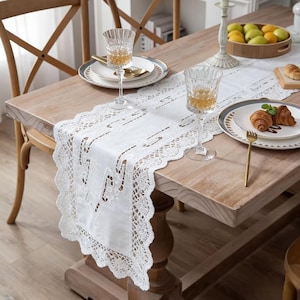 May include: A white lace table runner with a scalloped edge is displayed on a wooden table. Two wine glasses, plates, and a gold fork are set on the table. A wooden chair is visible in the background.