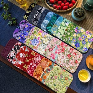 Op de afbeelding: Een verzameling kleurrijke placemats met patronen. De placemats hebben bloemen-, geometrische en door de natuur geïnspireerde ontwerpen in verschillende kleuren, waaronder rood, blauw, groen en paars. Ze zijn gerangschikt op een donkere ondergrond, met theekopjes en een fruitmand op de achtergrond.