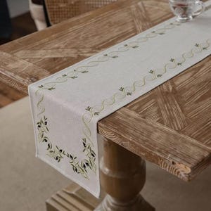 Può includere: Runner da tavolo in lino beige con ricamo di rami d'ulivo. Il runner presenta un motivo ripetuto di rami d'ulivo lungo i bordi e un motivo dettagliato di rami d'ulivo agli angoli. È esposto su un tavolo di legno.