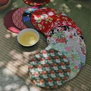 Könnte beinhalten: Eine Sammlung bunter, runder Stoffuntersetzer mit japanisch inspirierten Blumen- und geometrischen Mustern. Eine kleine weiße Teetasse mit Tee steht zwischen den Untersetzern. Die Untersetzer sind auf einer gewebten Matte angeordnet.