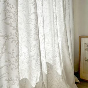 May include: White curtains with a subtle floral pattern. The fabric drapes in soft folds, creating a sense of elegance. The design features delicate botanical elements, adding a touch of nature to the room. The curtains are a neutral color, suitable for various interior styles.