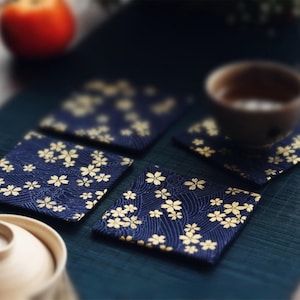Könnte beinhalten: Vier quadratische Untersetzer in Marineblau mit einem goldenen Blumenmuster. Die Untersetzer sind auf einer dunkelgrünen Oberfläche angeordnet, mit einem unscharfen Hintergrund, der eine Teetasse und eine Frucht umfasst.