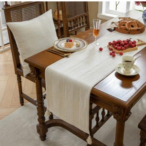 May include: A dining table set with a cream-coloured table runner, a matching cushion on a wooden chair, and a plate of cake. The table is set with a glass of wine, a teacup, and a basket of bagels. The runner has tassels.
