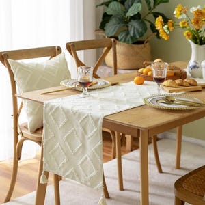 May include: A dining table set for a meal, with a cream-coloured table runner featuring a diamond pattern and tassels. The table is set with plates, glasses, and gold cutlery. A matching cushion rests on a wooden chair. A vase of yellow flowers and a potted plant are in the background.