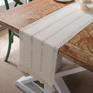 May include: Cream-colored table runner with a woven design and fringe detail. The runner is placed on a wooden table, adding a touch of texture and style to the dining or living space. The neutral color complements various decor styles.