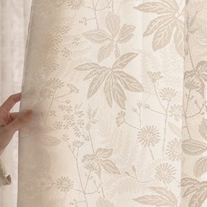 May include: Cream-coloured lace curtain with a repeating floral pattern. The design features large leaves, small flowers, and delicate stems. The curtain is held open, revealing the intricate details of the fabric.