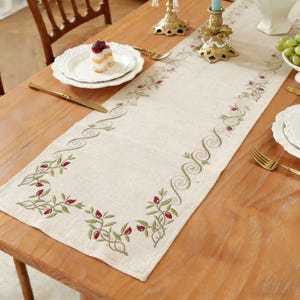 Puede incluir: Camino de mesa color crema con bordados de hojas verdes y bayas rojas. El camino de mesa rectangular está colocado sobre una mesa de madera. Remolinos decorativos y patrones florales adornan los bordes.