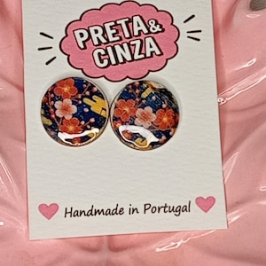 Pode incluir: Dois brincos circulares com fundo azul escuro e um padrão floral vermelho e branco. Os brincos estão expostos num cartão branco com o texto PRETA & CINZA numa forma de nuvem rosa e o texto Handmade in Portugal.