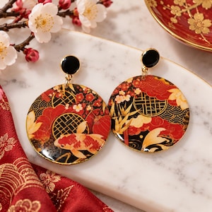 Pode incluir: Um par de brincos redondos com um padrão em preto, vermelho e dourado com peixes koi, bambu e motivos de nuvens. Cada brinco tem um pequeno pino circular preto no topo, ligado por um elo de metal dourado ao pendente circular maior.