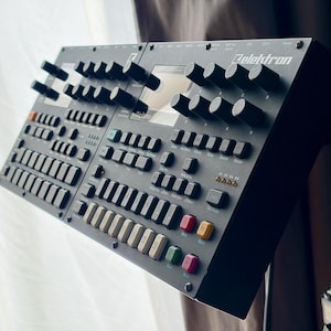 Puede incluir: Una caja de ritmos y sampler Elektron Digitakt de color gris oscuro con una pantalla central, numerosos mandos giratorios negros, una cuadrícula de botones cuadrados de goma y una fila de botones de colores amarillo, verde y rosa, con texto blanco.