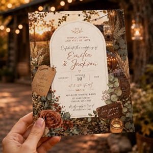 May include: A wedding invitation card featuring an arched cream-colored center with the text 'Celebrate the wedding of Emilia & Jackson', 'October 10th 2026', and 'Willow Grove Barn'. It is decorated with brown roses, eucalyptus, and tags.