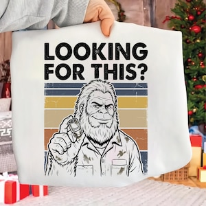 以下が含まれることがあります： 10mmのソケットレンチを持っているビッグフットのグラフィックが描かれた白いTシャツ。「LOOKING FOR THIS?」の文字がビッグフットのイラストの上にあります。背景には、茶色、ベージュ、青色の横縞模様があります。