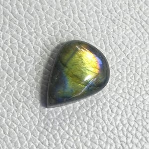 Puede incluir: Una piedra preciosa de labradorita pulida en forma de lágrima. La piedra muestra una mezcla de colores, incluyendo verde, amarillo y azul, con un efecto brillante e iridiscente. La piedra preciosa está sobre una superficie blanca texturizada.