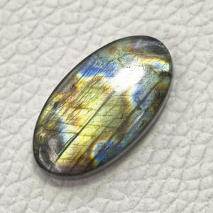 May include: An oval-shaped labradorite gemstone with a polished surface. The stone displays iridescent colors of blue, yellow, and green, with a dark gray base. The gemstone is set against a white textured background.