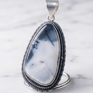 May include: A teardrop-shaped pendant featuring a white stone with dark gray dendritic inclusions. The stone is set in a silver frame with a decorative scalloped edge and a twisted rope border, topped with a polished silver bail for hanging.