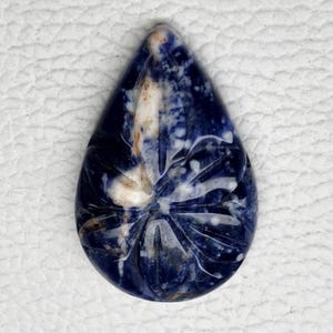 May include: A teardrop-shaped gemstone with a carved floral design. The stone features a deep blue hue with white and tan inclusions. The carved design is visible on the surface of the stone, adding texture and detail. The gemstone is set against a white background.