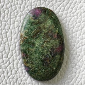 Puede incluir: Una piedra preciosa en forma de cabujón ovalado con un color base verde intenso y prominentes inclusiones de depósitos minerales magenta y rojo oscuro en toda la superficie, mostrando una textura pulida y lisa con variaciones naturales.