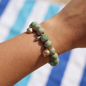 Peut inclure: Un bracelet composé de perles rondes vert marbré à la finition lisse. Le bracelet est agrémenté de petites breloques métalliques dorées en forme de coquillages et d'un coquillage conique pointu, espacées régulièrement entre les perles.