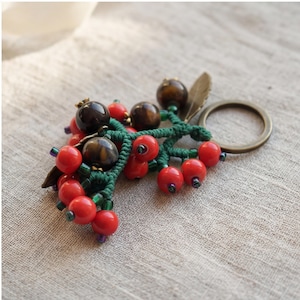 May include: A handcrafted keyring with a cluster of red and brown beads, joined by green macrame. The keyring includes a bronze-coloured ring and a small bronze-coloured leaf charm. The beads are arranged to resemble berries.