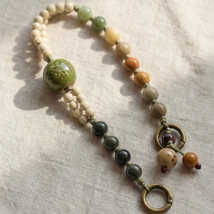May include: A beaded keychain with a variety of earth-toned beads. The keychain features a large green ceramic bead, cream-colored beads, and a gradient of brown, yellow, and green beads. It has a bronze-colored clasp and a small tassel.