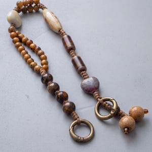 May include: A beaded lanyard featuring a variety of brown, tan, and cream-colored beads in spherical, cylindrical, and heart shapes. The beads are strung on a tan cord and finished with two circular bronze-toned metal lobster claw clasps.