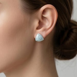以下が含まれることがあります： 明るい青色の三角形スタッドイヤリングのクローズアップ。イヤリングはテクスチャ加工された表面を持ち、耳と髪の毛を背景に配置されています。イヤリングはシンプルでエレガントなジュエリーです。