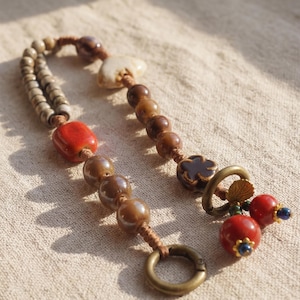 May include: A handcrafted beaded keychain featuring a variety of beads in shades of brown, red, and beige. The keychain includes a large red bead, a flower-shaped bead, and a metal ring for attaching keys.
