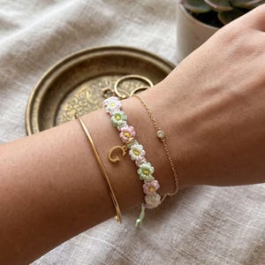 Puede incluir: Un conjunto de tres pulseras compuesto por un brazalete dorado liso, una pulsera tejida con flores en tonos pastel rosa, verde y blanco con un dije dorado de la letra G, y una fina cadena dorada con una pequeña piedra transparente engastada.