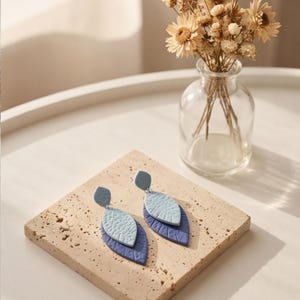 May include: A pair of handmade earrings with a layered leaf design. The earrings feature a light blue top, a textured light blue middle layer, and a dark blue bottom layer. They are displayed on a square, textured, beige stone.
