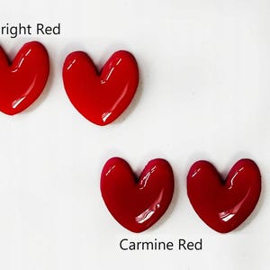 May include: Four glossy heart-shaped objects arranged in two pairs. The top pair is labeled Bright Red, featuring a vibrant, saturated red hue. The bottom pair is labeled Carmine Red, displaying a slightly deeper, richer shade of red.