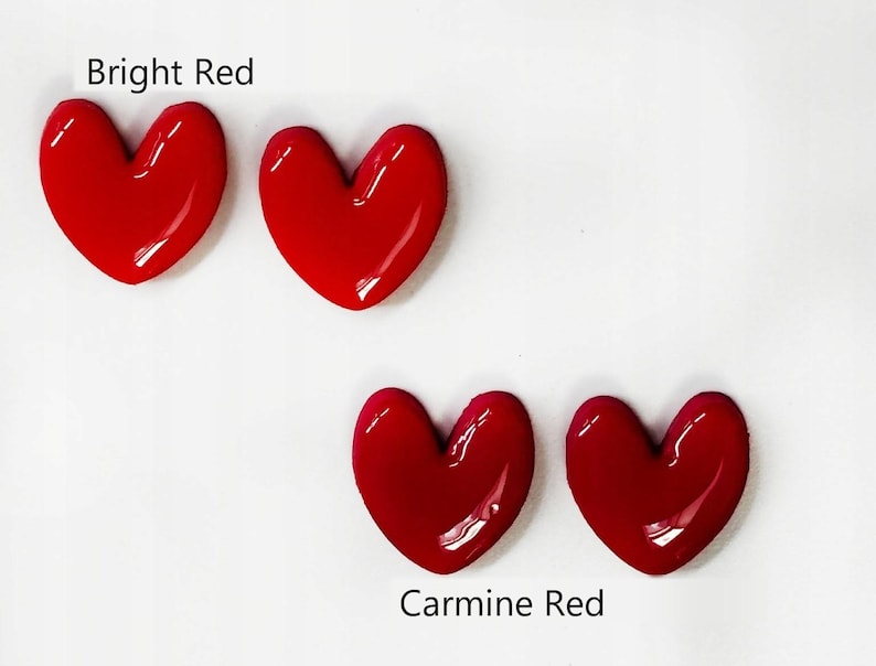 May include: Four glossy heart-shaped objects arranged in two pairs. The top pair is labeled Bright Red, featuring a vibrant, saturated red hue. The bottom pair is labeled Carmine Red, displaying a slightly deeper, richer shade of red.
