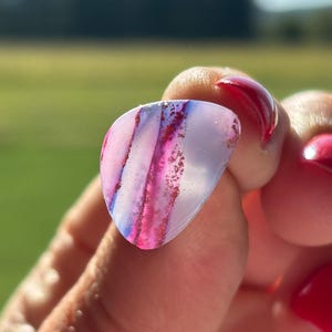 May include: A translucent, teardrop-shaped guitar pick featuring a smooth, polished surface with distinct vertical bands of vibrant magenta and deep purple, accented by fine, sparkling red glitter particles embedded within the material.
