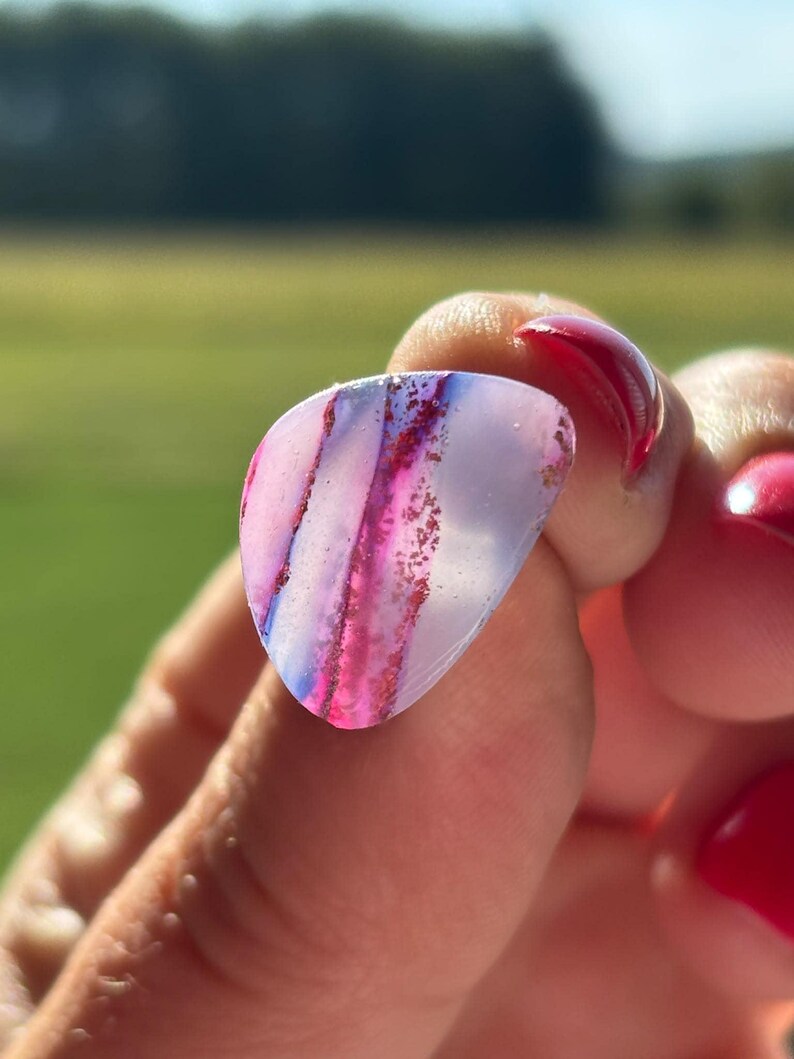 May include: A translucent, teardrop-shaped guitar pick featuring a smooth, polished surface with distinct vertical bands of vibrant magenta and deep purple, accented by fine, sparkling red glitter particles embedded within the material.